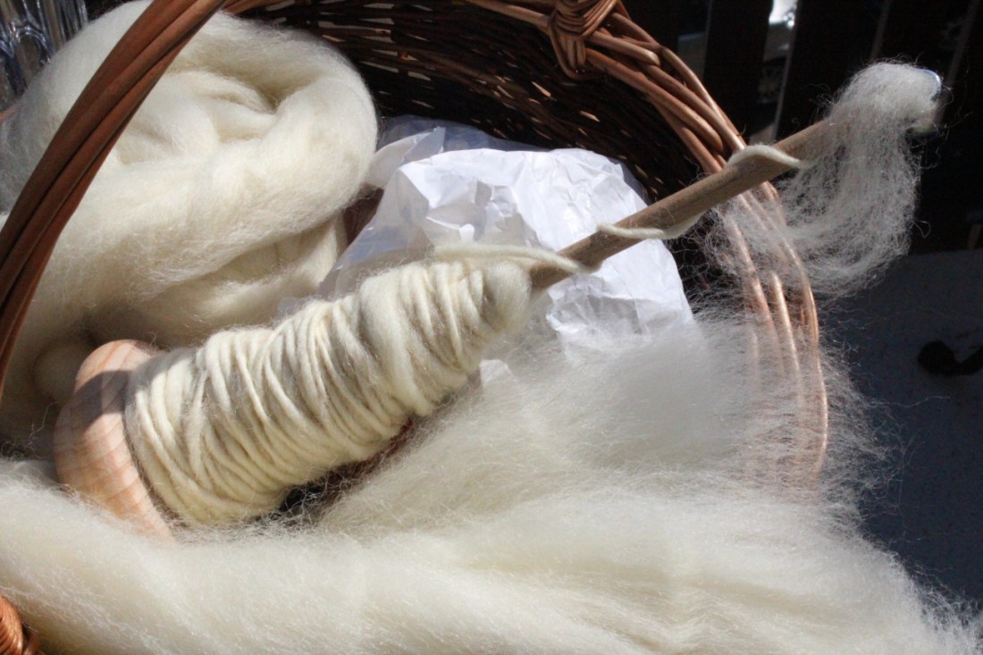 Ein Korb, in dem sich eine Papiertüte, ein Bausch Fasern aus cremeweißer Eiderwolle und eine Handspindel befindet, die zur Hälfte mit Eiderwolle vollgesponnen ist.