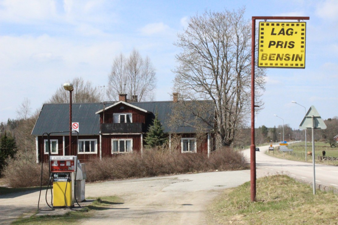 Verlassene Selbstbedienungstankstelle vor einem schwedenroten Familienhaus. Zwei freistehende Zapfsäulen ohne Dach, am straßenrand ein Schild auf dem auf Schwedisch "Niedrigpreisbenzin" steht.
