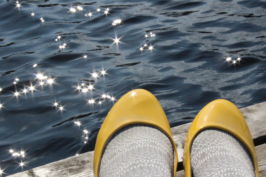 Steg am Wasser,. darauf die Spitzen von gelben Lackschuhen, in denen hellgraue Strumpfhosenbeine stecken, die Sonne reflektiert Sterne auf das dunkle Wasser.