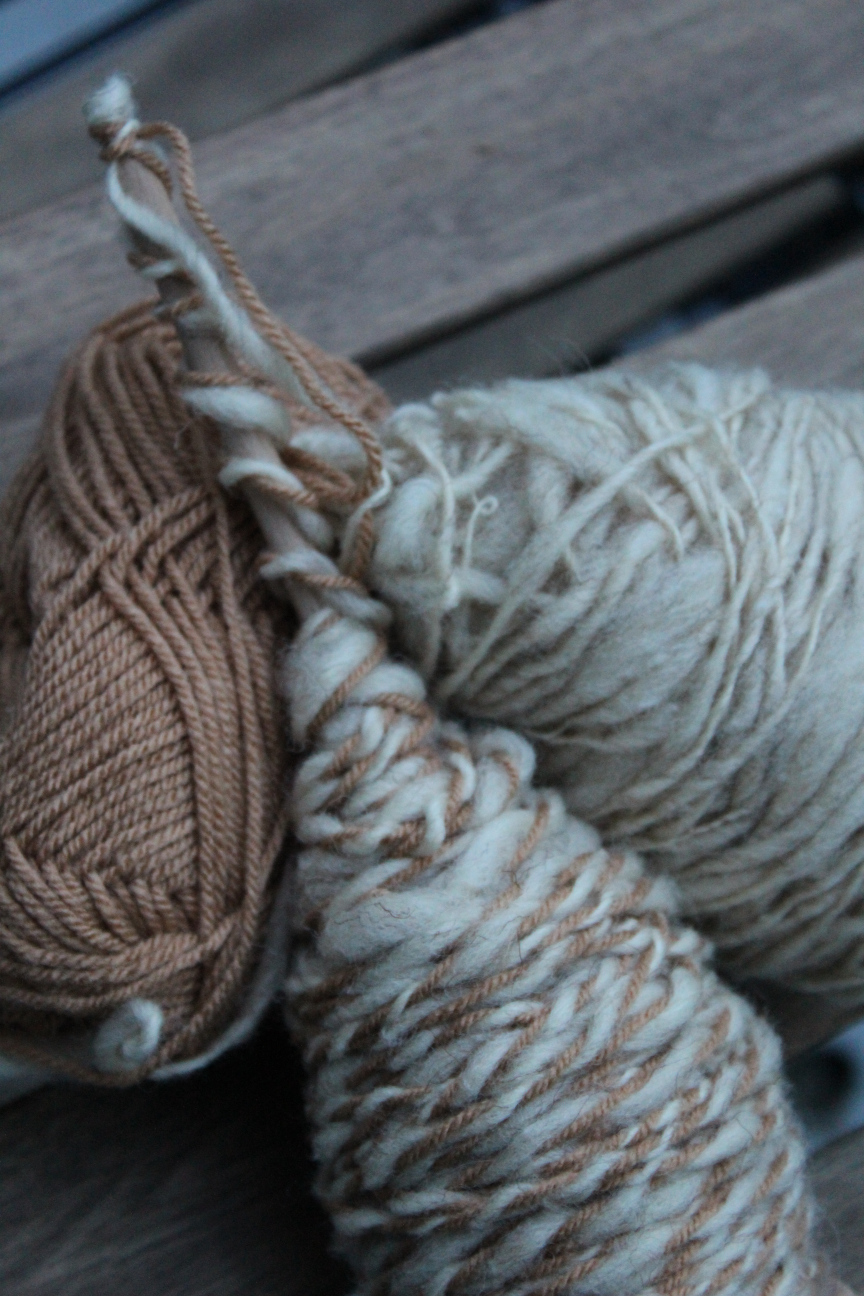 Mit verzwirntem Garn gefüllte Spindel liegt zwischen einem hellbraunen Polyacrylgarn und frisch gesponnener und unsauber aufgewickelter Wolle vom Coburger Fuchs auf einem Balkontischchen rum. Wenn man genau hinguckt, sieht es so aus, als seien bei dem verzwirnten Garn die Stellen, an denen man das hellbraune Garn sieht, kleine Maden anstelle von Garn. Bäh.