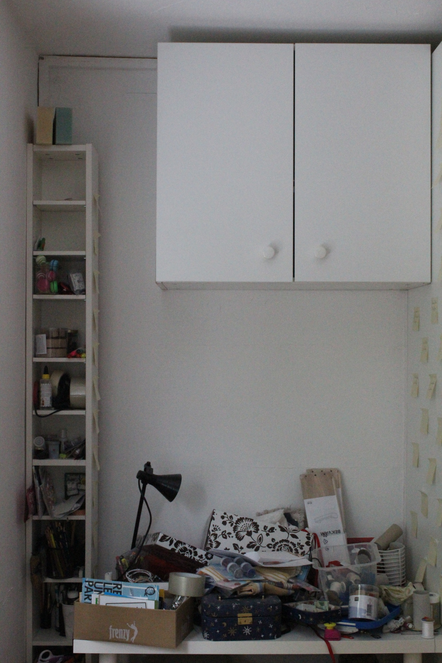 Ein weißer Schreibtisch, vollgestapelt mit Zeug und Kram und Kisten, Bastelsachen, eine Lampe, Postkarten und Glitzertuben. Links davon ein weißes Ikea-CD-Regal, eion bisschen aufgeräumter, darin Kleber und Scheren und Einmachgläser mit Krimskrams. Oben drüber ein weißer Schrank an der Wand, beide Türen verschlossen, endlich was, das ordentlich aussieht.