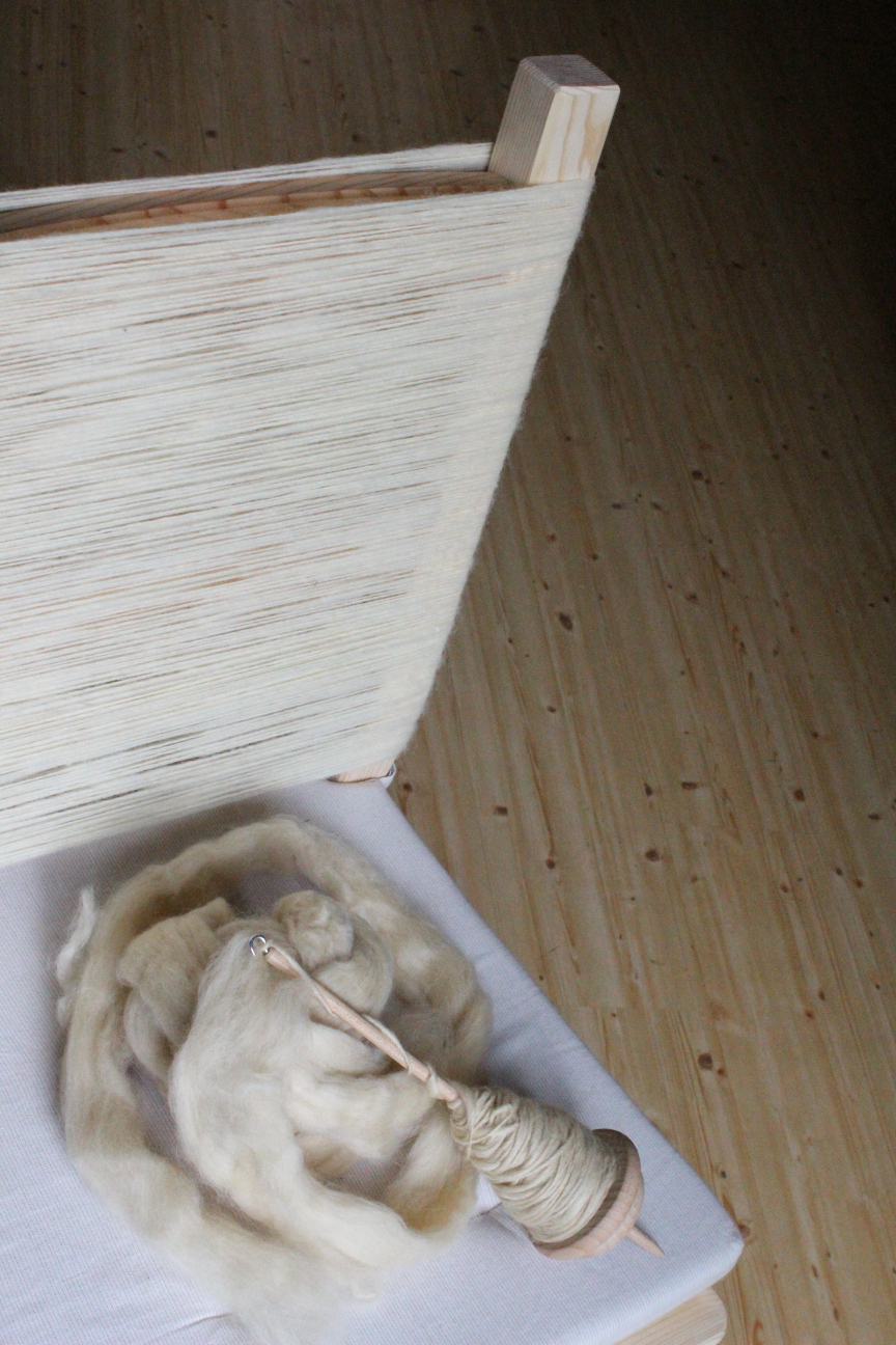 Halber Ausschnitt von einem Stuhl, dessen Lehne komplett mit feinem weißem Garn aufgewickelt ist. Auf dem Sitz liegt eine Handspindel mit etwas gelberen Fasern, die schon angesponnen sind.