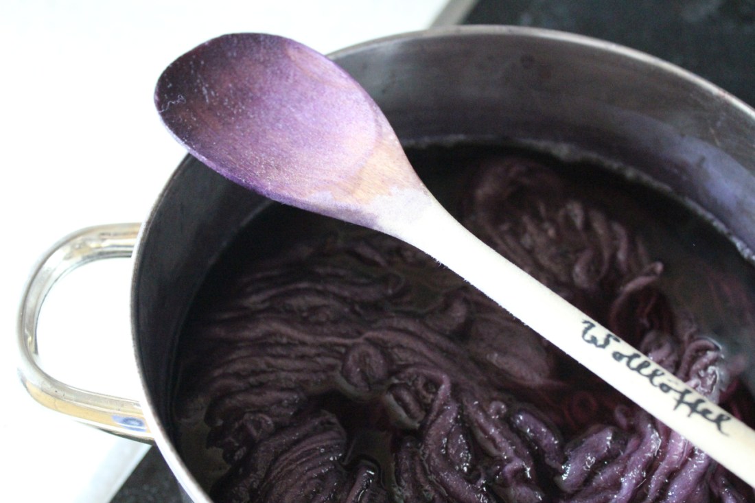 Lila Wolle im Farbbad im Topf. Der Löffel, auf dem "Wollöffel" steht, hat einen lila Spitze.