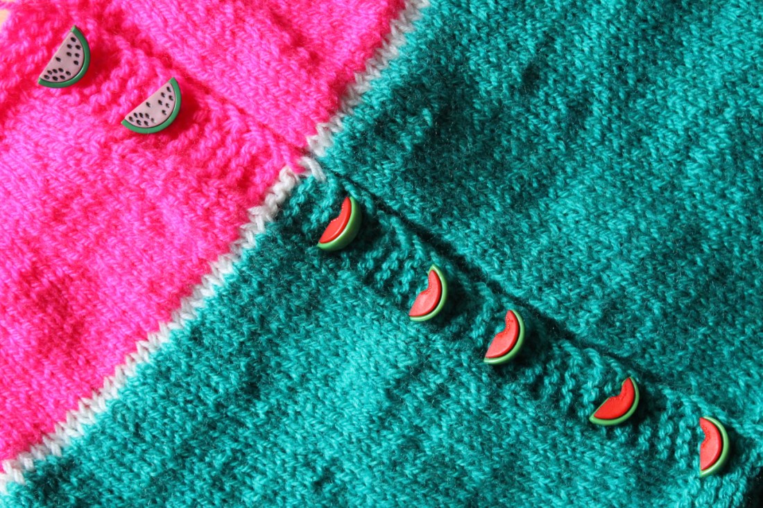Knopfleiste einer Strickjacke, die zu einem drittel pink, zu zwei dritteln grün ist. Auf der Knopfleiste liegen wassermelonenförmige Knöpfe, auf der grünen Wolle mit rotem Fruchtfleisch, auf der pinken Wolle mit rosa Fruchtfleisch.