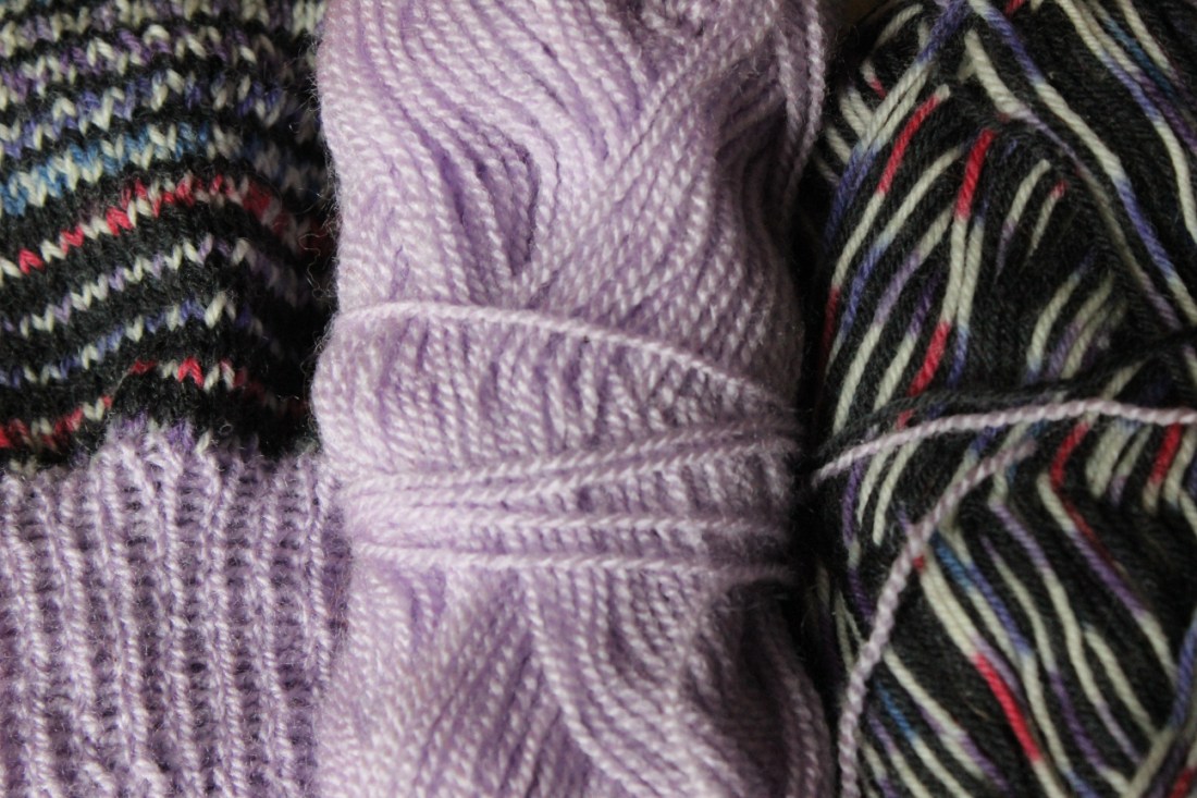 Zwei Garnknäuel, eines helllila, das andere schwarz mit weiß und lilarotblau gemischt liegen neben einem Strickstück, in dem beide Knäuel zu gleichen Teilen verstrickt sind.