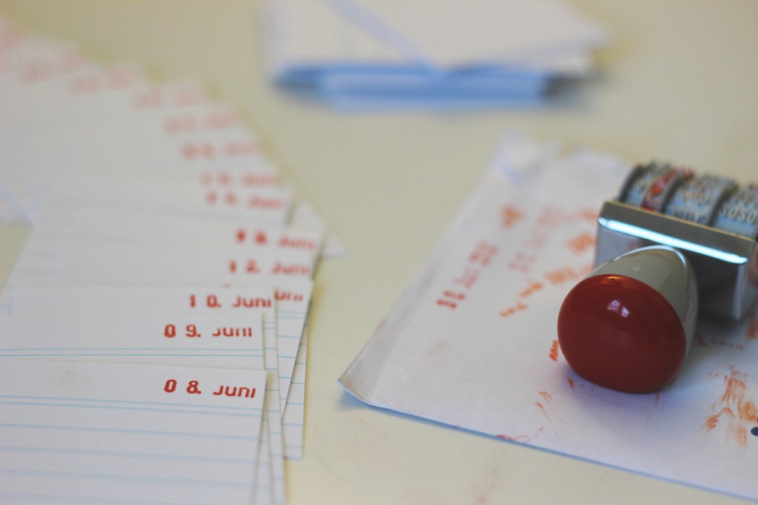 Viele Karteikarten, auf denen je ein Datum oben rechts hingestempelt ist. Im Vordergrund der 8. Juni.Rechts daneben ein Datumsstempel auf einem verschmierten Papierumschlag. hinten noch ein Stapel Karteikarten.