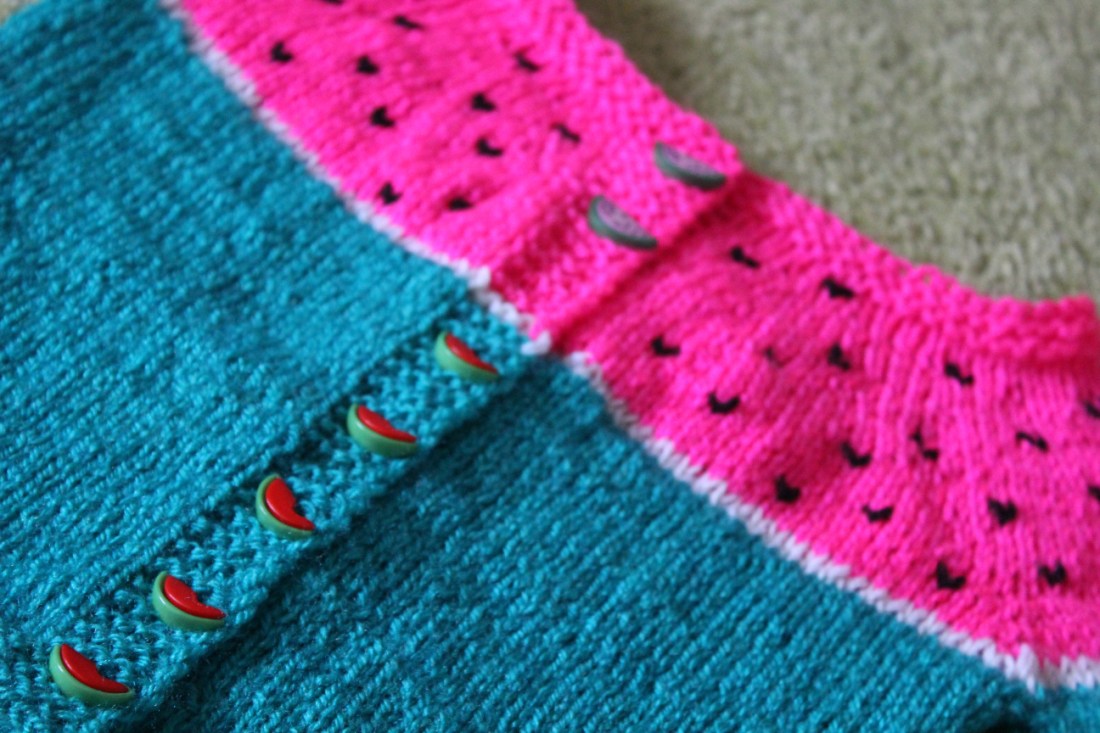 Nahansicht der Knopfleiste, einer Wassermelonenjacke, am Kragen viel pinke Wolle, von der Brust an grüne Wolle. Auf der grünen Wolle rote Wassermelonenknöpfe, in denen ein Biss drin ist, auf der pinken Wolle rosa Wassermelonenknöpfe mit schwarzen Kernflecken drin.  In die pinke Wolle sind Kerne mit schwarzer Wolle gestickt.
