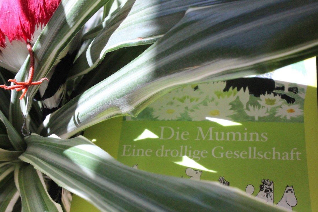 Das Buch "Die Mumins, eine drolliige Gesellschaft" von Tove Jansson hinter ein paar Zimmerpflanzenblättern, auf denen ein Vogel sitzt, von dem man nur das Plastikbeinchen im Licht sieht.