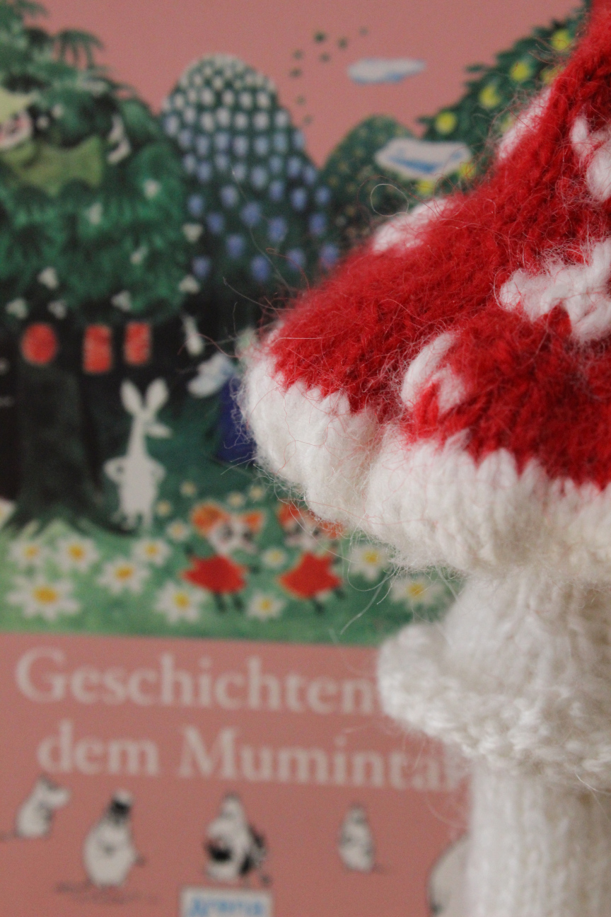 Das Cover des Buches "Geschichten aus dem Mumintal" von Tove Jansson, leiucht verschwommen hinter einem selbstgestrickten Fliegenpilz.