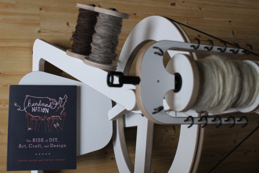 Ein weißes Spinnrad, auf dessen Pedal das Buch "Handmade Nation - The Rise of DIY, Art, Craft, and Design" von Faythe Levine und Cortney Heimerl liegt.