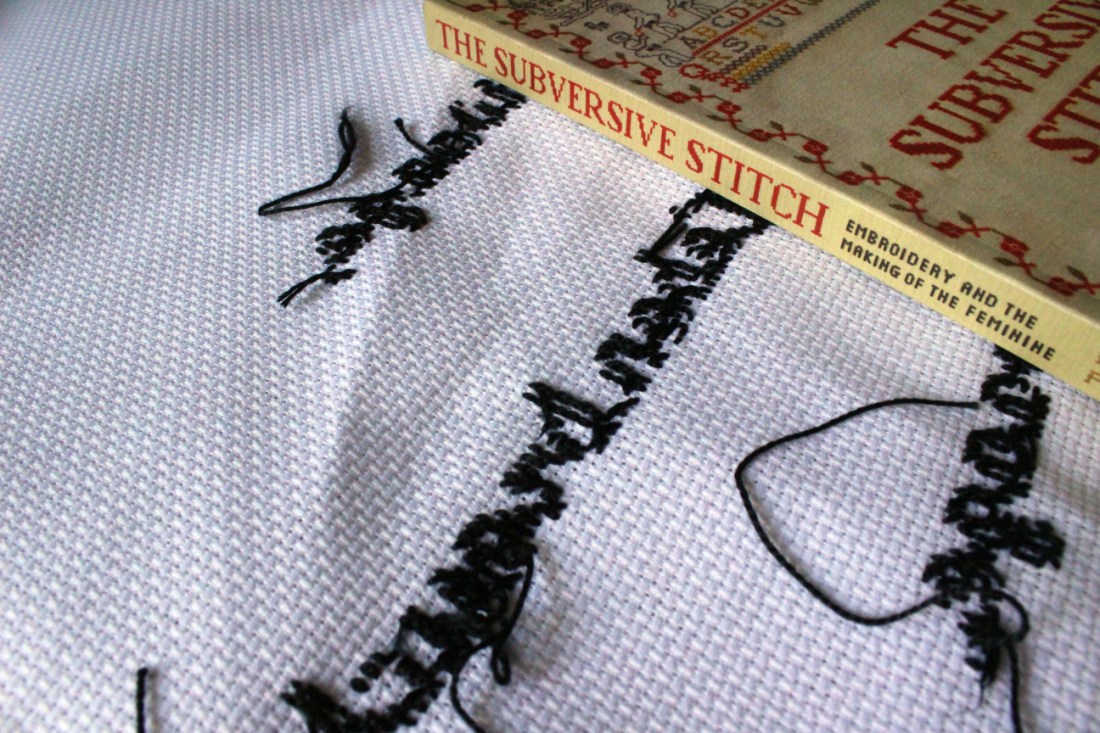 Auf der Rückseite eines weißen mit schwarzer Schrift bestickten Straminstoffes liegt ein beiges Buch im rechten oberen Eck des Bildes, so dass man nur den Buchrücken gut sieht. Es ist das Buch "The Subversive Stitch - Embroidery and the Making of the Feminine" von Rozsika Parker