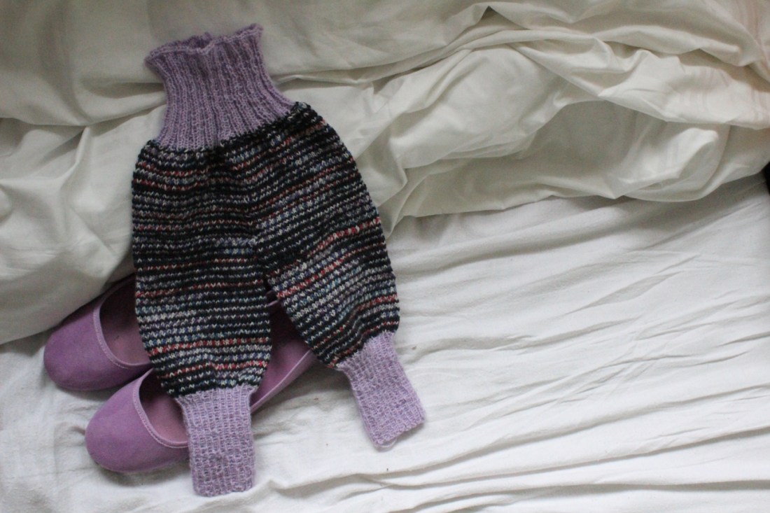 Selbstgestrickte Krembelhose mit lila Fuß- und Bauchteil und dunkelgestreiften Beinen, die auf zwei lila Ballerinas in einem weißbezogenen Bett liegt.