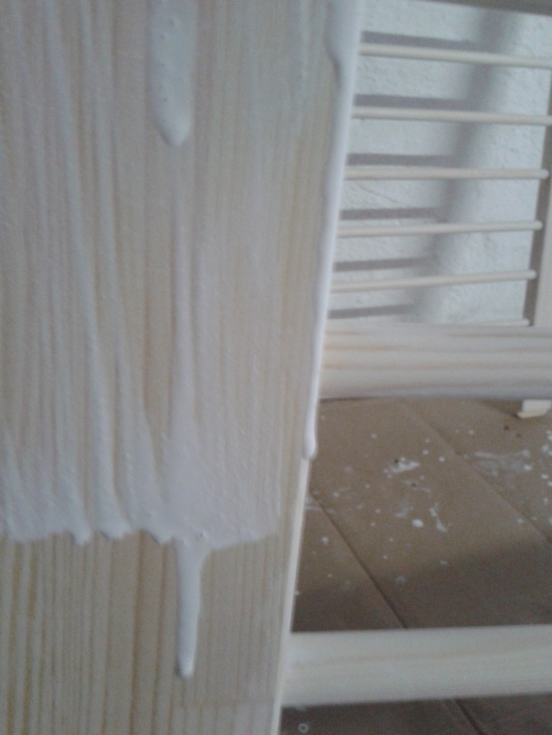 Mit weißer Holzlasur bestichene Holzplatte, von der die Lasur runtertropft, vor einer bereits gestrichenen Sprossenwand.