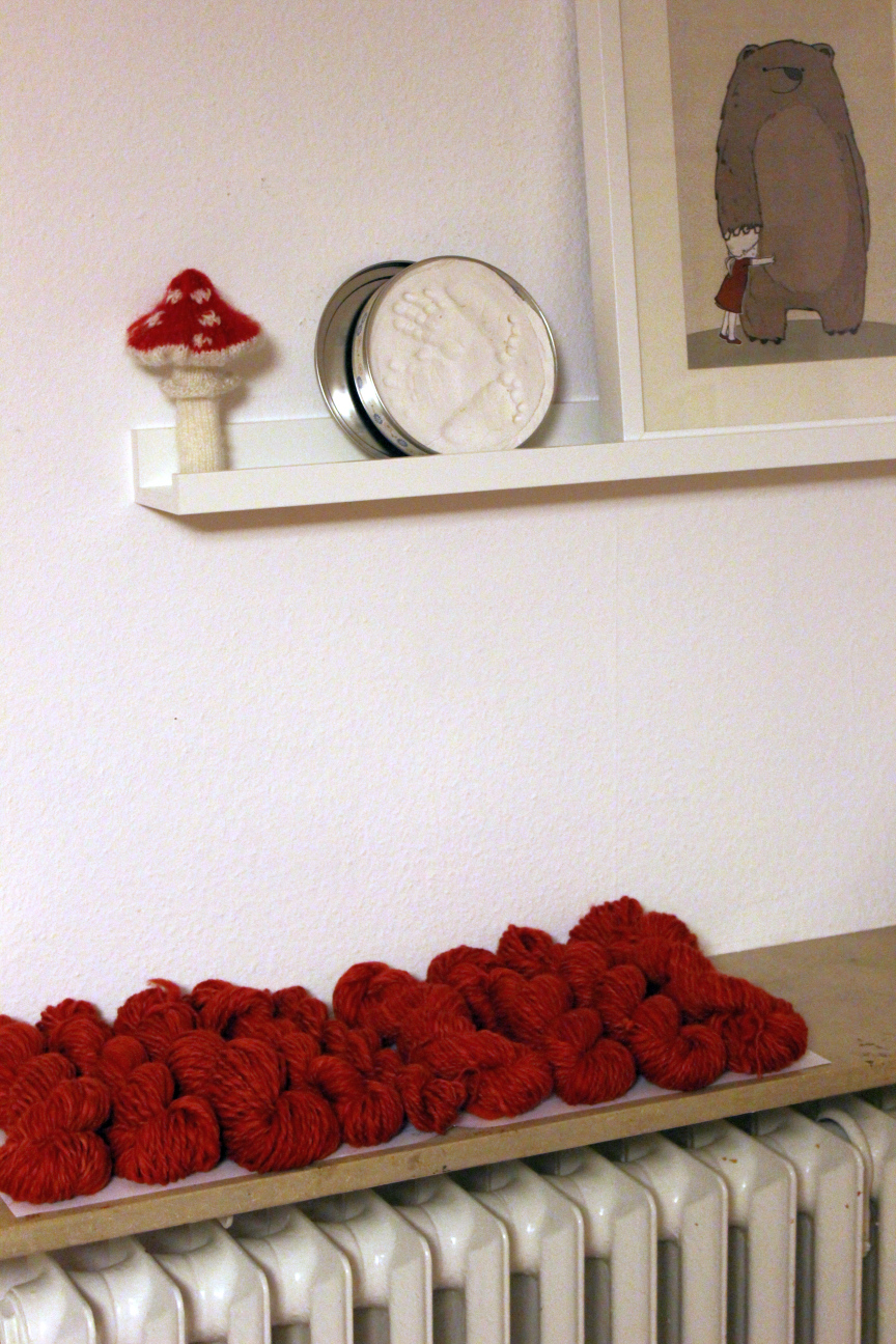 Nein Stränge selbst gesponnene Wolle nebeneinander aufgereiht, darüber an der Wand ein kleines Regal, auf dem ein selbstgestrickter Fliegenpilz, ein Gipsabdruck mit Baby-Händen- und Füßen und einer Kinderzimmerillustration von Martina Hoffmann mit einem Bären, der den Kopf eines rotgekleideten Mädchens tätschelt.