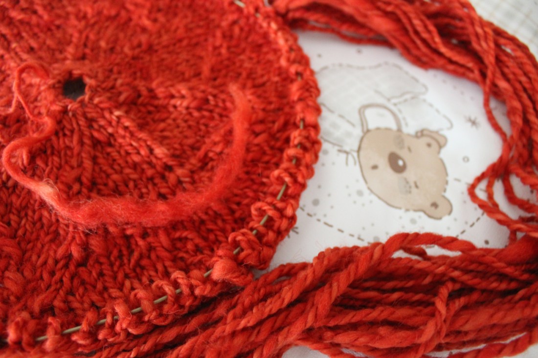 andere angestrickte Babydecke aus dem gleichen Garn. liegt auf einer Wickelauflage, auf der ein schlafendes Bärchen abgebildet ist.