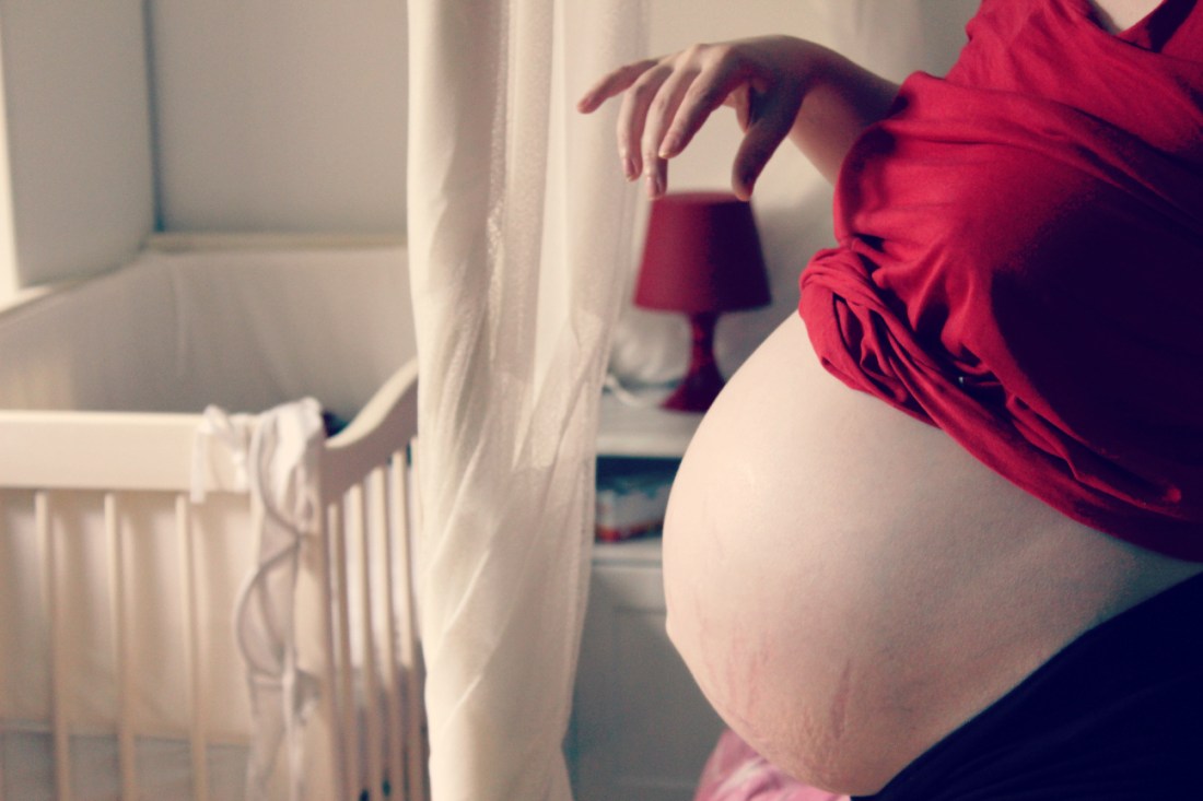 Links im Hintergrund ein weiß lasiertes Baby-Bettchen, rechts im Vordergrund mein nackte Bauch, das rote T-Shirt hochgezogen, meine rechte Hand spielt mit einer roten Lampe im Hintergrund