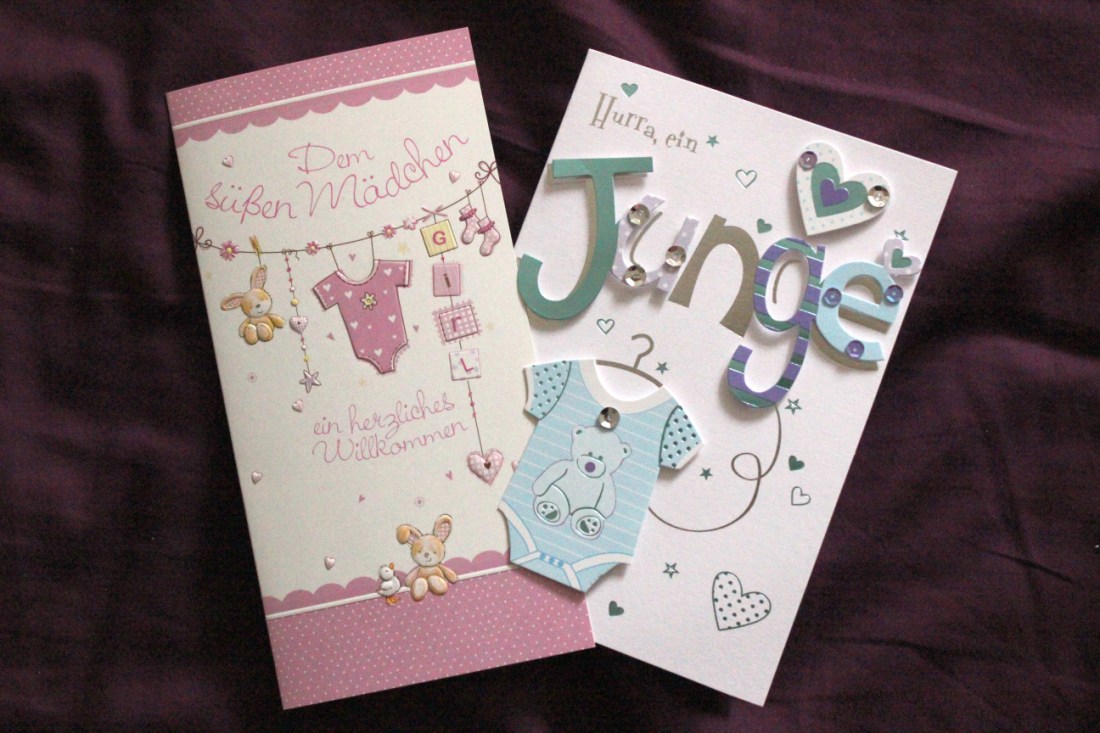 Zwei Glückwunschkarten, auf deren Titelseite in rosa Dekoration Glückwünsche zum Mädchen und in hellblauer Dekoration Glückwünsche zum Jungen stehen.