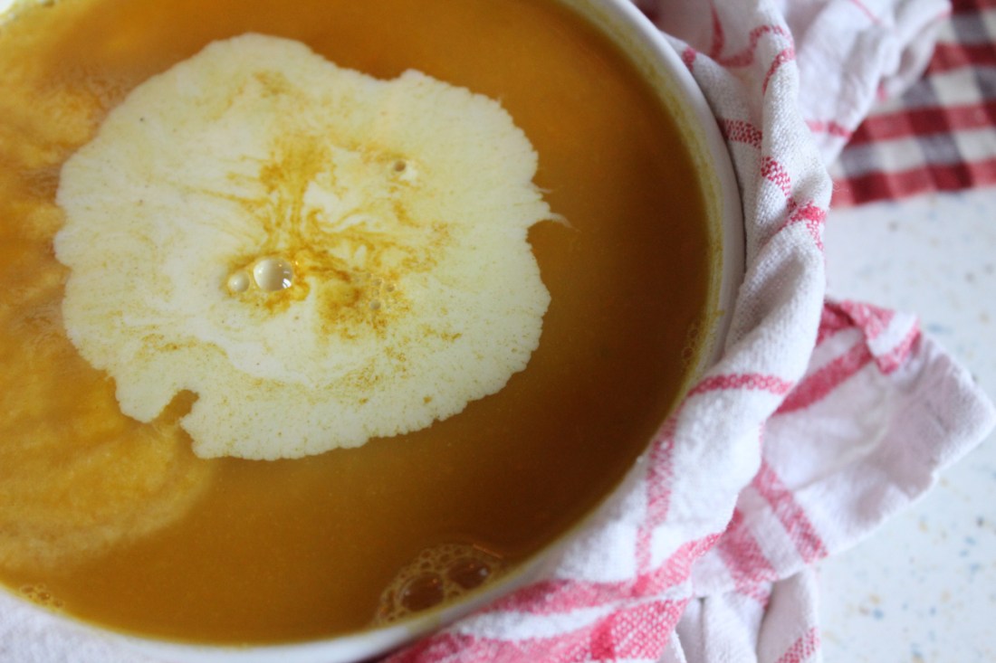 Voller Teller Kürbissuppe mit Klecks Sahne drin, eingeschlungen in ein Geschirrhandtuch mit rosa Karos.