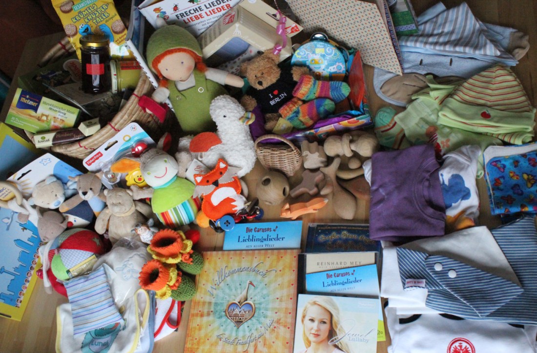 Foto mit fast allen Geschenken zur Geburt des Babys, ausgebreitet auf einem Tisch, Bücher, CDs, Kuscheltiere, Spucktücher, Rasseln, Kleidung, Schuhe, Puppen, Spielfiguren, Essen und mehr.