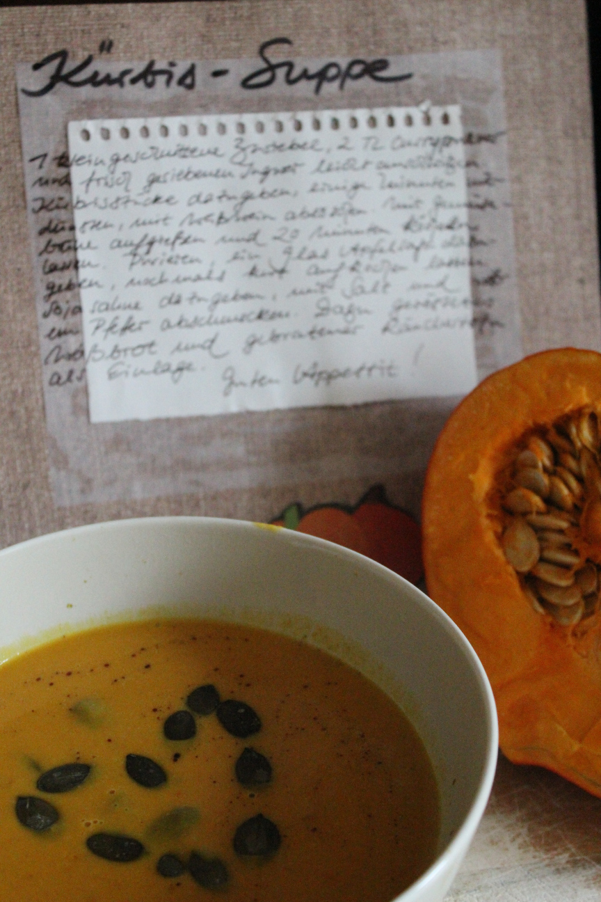 Die fertige Suppe und die übriggebliebene Kürbishälfte vor der aufgeschlagenen Rezeptseite; in der suppe schwimmen Kürbiskerne in Herzformation.