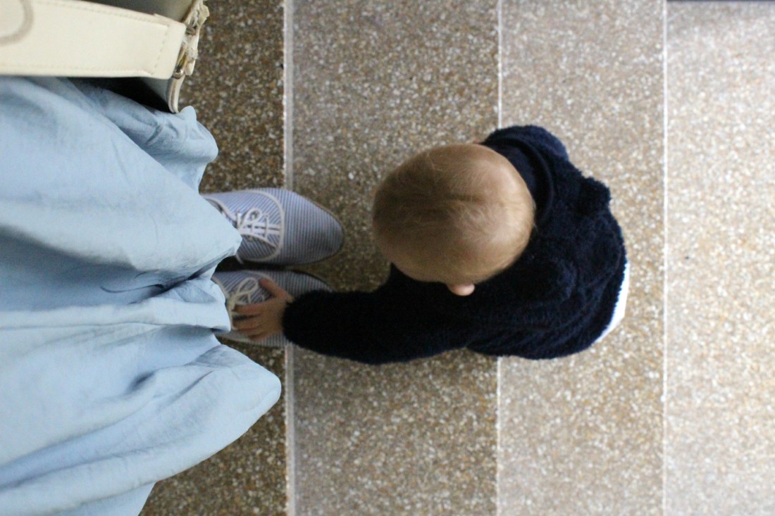 Vier Treppenstufen, von oben draufgeguckt: auf der obersten steht eine erwachsene Person, von ihr sind nur zwei blei-weiß-gestreifte Stoffstchuhe, ein taubenblauer Rock und eine weiße Kunstledertasche zu sehen. Die Person guckt in Richtung trepperunter. Zwei Stufen unter ihr steht ein Baby in einem dunkelblauen Teddyfelljäckchen mit Kapuze. Das Baby ist die Stufen von unten nach oben hochgeklettert und fasst mit der linken Hand den rechten Schuh der erwachsenen Person an. 
