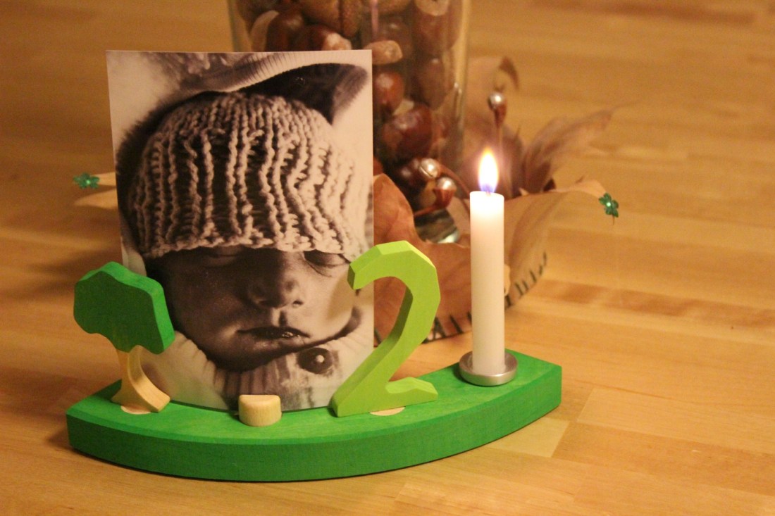 Vor einer mit Kastanien gefüllten Vase, die mit einer Krone aus getrockneten ahornblättern umfasst sind, an deren Spitzel grüne Pailletten und silberne Miniglöckchen kleben, steht ein grünes Viertel aus einem Holzgeburtstagsring, in dem eine brennende weiße Kerne, eine grüne 2, ein kleiner Baum und ein Schwarz-Weiß-foto mit einem Babay, das die Augen geschlossen und schwarze Lippen hat, stecken. Es trägt eine Wollmütze, halb über die Augen gezogen.