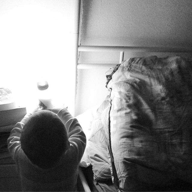 Das Kind steht neben dem Bett, von hinten fotografiert und hebt die Milchflasche mit beiden Arme hoch. Durch sie strahlt das Licht der Nachttischlampe.